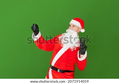 A Caucasian man dressed as Santa Claus is taking a picture of himself with a cell phone. With the other hand he is making the victory gesture. The background is green.