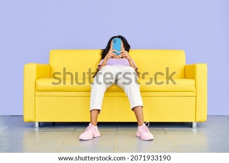 Funny photo of woman types text message on cell phone, enjoys online communication lying on yellow sofa