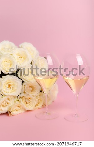 Beautiful romantic composition with white roses and two glasses of white wine on pink background. French white wine for romantic dinner. Holiday card with flowers and wine. Happy Valentine's Day