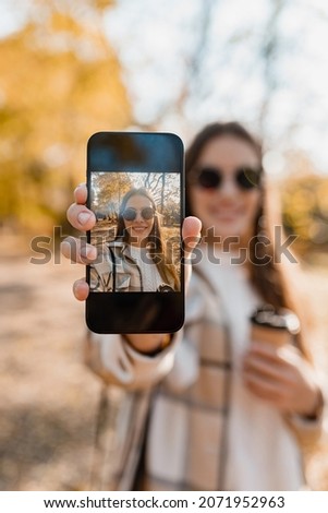 attractive young woman walking in autumn park taking selfie pictures using smartphone, wearing checkered coat, sunglasses, happy mood, fashion style trend