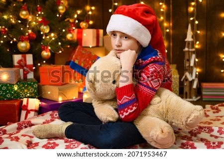 Child girl posing in new year or christmas decoration. Festive lights and lots of gifts, an elegant Christmas tree with toys. The girl is wearing a red sweater and a Santa hat, she playing with bear