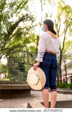 young latin tourist woman, standing in a park enjoying the afternoon, with a hat in her left hand, wearing a protective mask, model not recognizable.