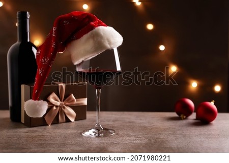 Glass of red wine with Santa Claus hat, bronze gift box and Christmas decorations on table. Christmas lights in the black background.