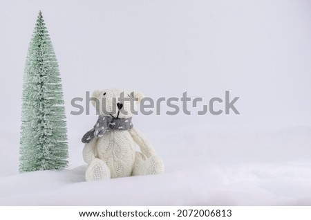 A white bear in the snow next to a snow-covered Christmas tree. A postcard with a knitted toy. Space for text.