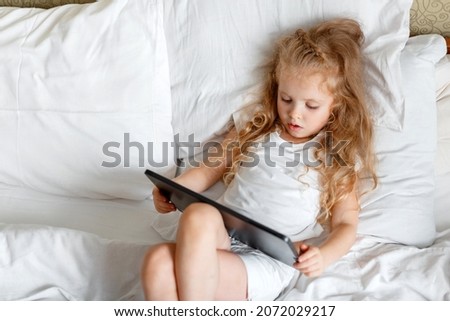 Serious attentive Caucasian Girl use Tablet for watching videos and cartoons. Beautiful Child girl Portrait using tablet Computer lying in bed. Little blond girl play game on tablet in bedroom at home