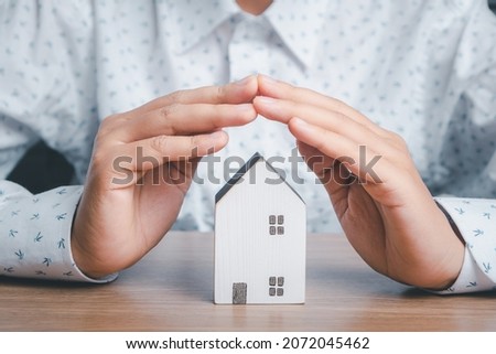 Mini house under hands in gesture of protection on wooden desk, homeless housing and home protecting insurance concept, international day of families, homeschooling