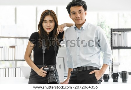 Portrait young male and female business partners in casual clothing standing in office looking at camera. Smart woman journalist hanging digital camera ready to go take pictures as reporter lifestyle.
