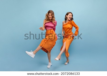 Full-length two beautiful cheerful caucasian young girls are dancing in photo studio. Blonde in pink top, skirt. Brown-haired woman in orange dress with bare shoulder in shoes.