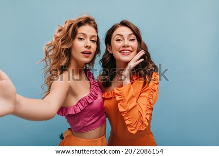 Close-up of two amazing caucasian girls make selfie on blue background. Pretty brunette and blonde with wavy hair in bright outfits. Leisure lifestyle and beauty concept.