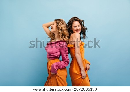 Two cute caucasian girls stand back to back laughing cheerfully in studio shot of blue color. Brunette is dressed in orange dress with bare shoulder. Blonde in pink blouse and orange pants. Royalty-Free Stock Photo #2072086190