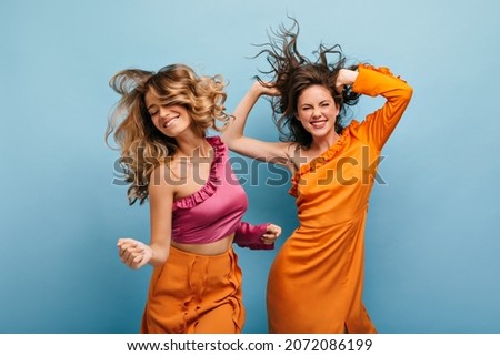 Cheerful showy caucasian young women fooling around on isolated blue background. Their wavy, loose hair flies in all directions. Girls are dressed in bright summer outfits. Royalty-Free Stock Photo #2072086199