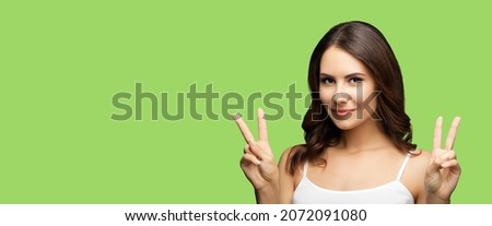 Photo of woman showing two fingers or victory hand sign gesture, isolated on light green background. Portrait of happy smiling gesturing brunette girl at studio. Wide horizontal banner composition