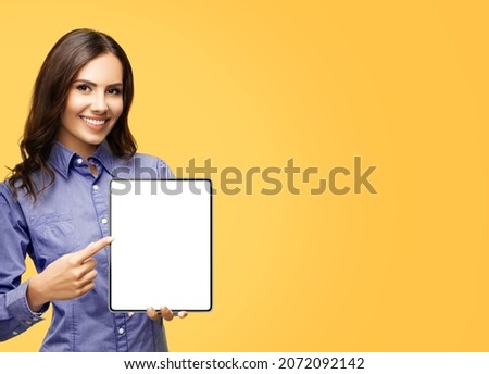 Happy smiling brunette businesswoman show blank tablet pc ipad touchpad monitor, with copy space area for some text, advertising slogan, isolated on yellow background. Business woman at studio image. 