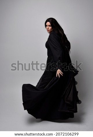 Full length portrait of dark haired girl wearing a witch black flowing gown and  fantasy cloak.   Standing pose with gestural movements, isolated on studio background. Royalty-Free Stock Photo #2072106299