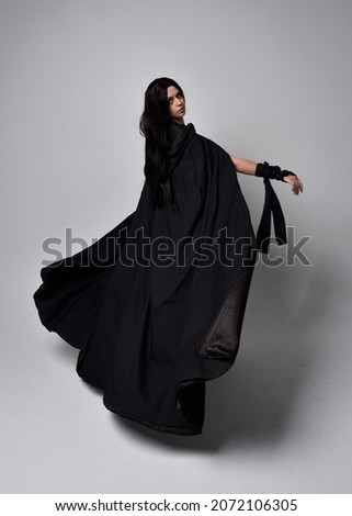 Full length portrait of dark haired girl wearing a witch black flowing gown and  fantasy cloak.   Standing pose with gestural movements, isolated on studio background.