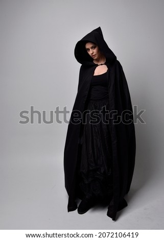 Full length portrait of dark haired girl wearing a witch black flowing gown and  fantasy cloak.   Standing pose with gestural movements, isolated on studio background. Royalty-Free Stock Photo #2072106419