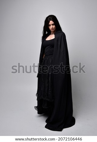 Full length portrait of dark haired girl wearing a witch black flowing gown and  fantasy cloak.   Standing pose with gestural movements, isolated on studio background.