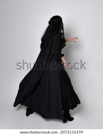 Full length portrait of dark haired girl wearing a fantasy witch black costume.   Standing pose with back to the camera and  gestural movements, isolated on studio background.
