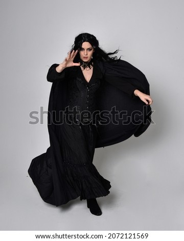 Full length portrait of dark haired girl wearing a fantasy witch black costume and flowing cloak.   Standing pose  gestural movements, isolated on studio background.