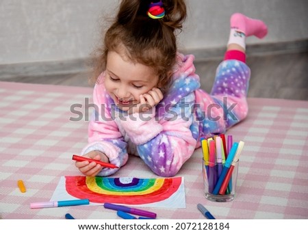A cute girl in rainbow clothes draws a rainbow with multi-colored markers. House. Childhood.