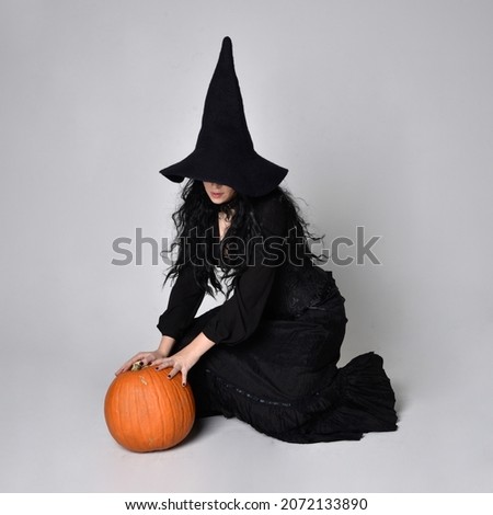 Full length portrait of dark haired girl wearing a fantasy witch black costume,  Sitting pose  with gestural movements, isolated on studio background.