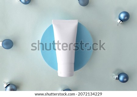 Mockup facial skincare white tube bottle on round pedestal display with soft light blue decorate with Christmas cyan baubles on plain grey paper background, Cosmetics decoration for festive season
