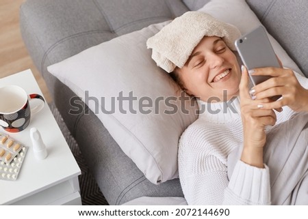Smiling happy woman using mobile phone while lying sick in bed, looking at device screen with positive expression, her husband trying to cheer up ill wife sending her funny messages. Royalty-Free Stock Photo #2072144690
