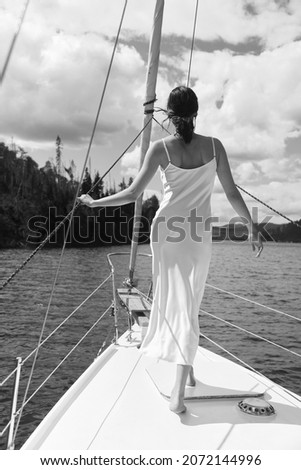 Gorgeous slim woman in the white silk dress on the white boat. Long legs, light skin. Brunette. Beautiful nature, clear water. Windy and sunny day. Vacation. Sea walk. Freedom. Black and white photo