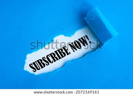 Text SUBSCRIBE NOW appearing behind torn blue paper. Top view.