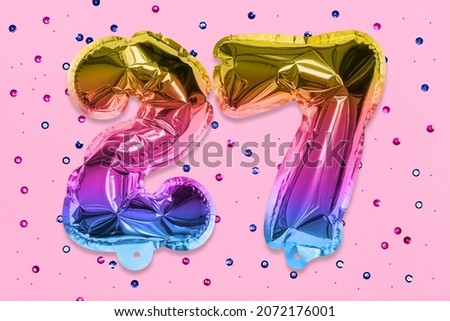 Rainbow foil balloon number, digit twenty seven on a pink background with sequins. Birthday greeting card with inscription 27. Top view. Numerical digit. Celebration event, template.