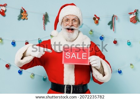 Old Santa Claus man 50s wears Christmas hat red suit clothes hold point on card sign with sale title text isolated on plain blue background studio. Happy New Year 2022 merry ho x-mas holiday concept