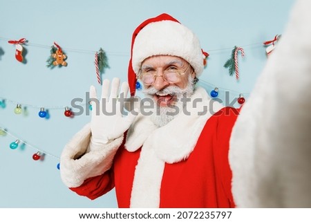 Close up old bearded Santa Claus man 50s in Christmas hat red suit do selfie shot pov on mobile phone waving hand isolated on plain blue background studio Happy New Year 2022 merry ho x-mas concept
