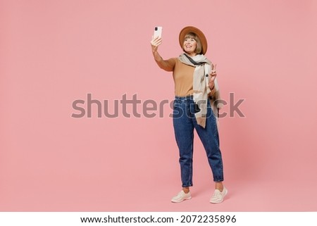Full size body length traveler tourist mature elderly woman 55 years old wears brown shirt hat scarf do selfie shot on mobile cell phone isolated on plain pastel light pink background studio portrait
