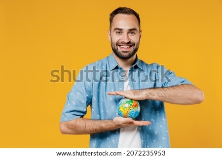 Young smiling happy cheerful friendly caucasian man 20s wearing blue shirt white t-shirt hold in palms Earth world globe isolated on plain yellow background studio portrait. People lifestyle concept