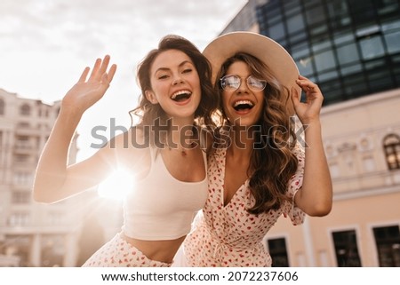 Bottom view of two cheerful beautiful caucasian young women posing for photo in sunbeams. On warm day, brunette and blonde are walking around city in white casual clothes. Royalty-Free Stock Photo #2072237606