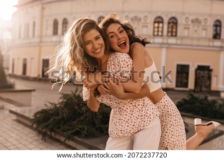 Caucasian young woman brunette hung on back of her friend against the open background of city. Girls are smiling broadly with their teeth at camera, dressed in white outfits. Royalty-Free Stock Photo #2072237720