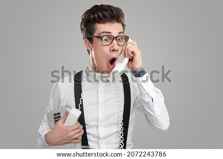 Dumbfounded male in eyeglasses with eyes wide open having phone call and feeling impressed by unexpected news standing on gray background