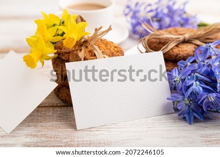 White paper business card mockup with oatmeal cookies with spring snowdrop flowers bluebells, narcissus and cup of coffee on white wooden background. side view, close up, still life. spring concept.