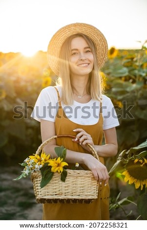 A woman is standing through a field of sunflowers with a basket of flowers in her hands. She is smiling and looking at the camera. Sunset in the background.