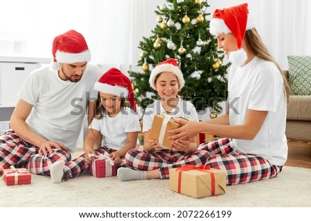 family, winter holidays and people concept - happy mother, father and two daughters in santa hats opening christmas gifts at home