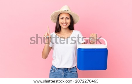 pretty hispanic woman smiling and looking friendly, showing number one and holding a portable refrigerator