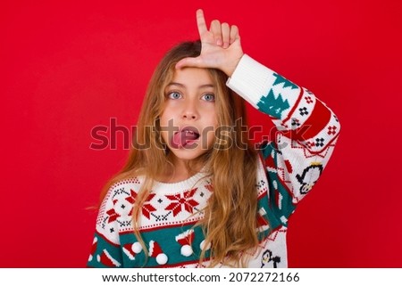 brunette kid girl in knitted sweater christmas over red background gestures with finger on forehead makes loser gesture makes fun of people shows tongue