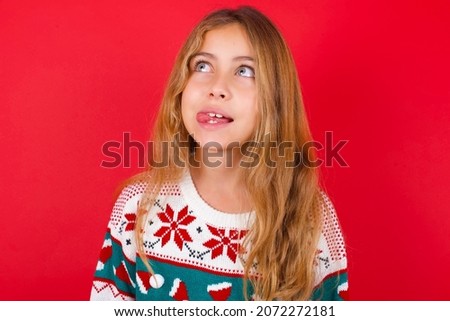 Funny brunette kid girl in knitted sweater christmas over red background makes grimace and crosses eyes plays fool has fun alone sticks out tongue.