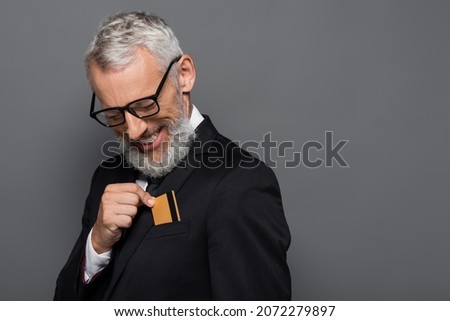 cheerful middle aged businessman in suit and glasses putting credit card in pocket isolated on grey