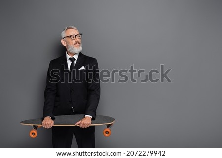 bearded middle aged businessman in suit and glasses holding longboard on grey