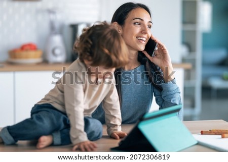 Shot of young smiling mother and her cute son watching cartoons on digital tablet while talking with smartphone in the kitchen at home.