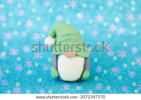 Christmas card figurine of a funny gnome with a beard and a big nose on a blue background and falling snowflakes.
