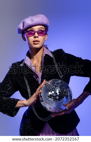 young woman in trendy sunglasses and blazer with animal print holding shiny disco ball on violet