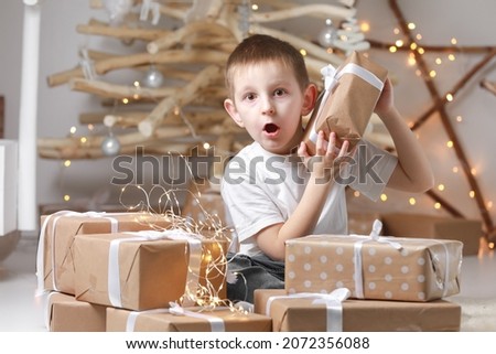 Holidays Concept. A cute little excited boy child is sitting by the wooden decorations Christmas tree and many gift boxes, holding a gift box. new Year's Eve and Christmas, waiting for a miracle.