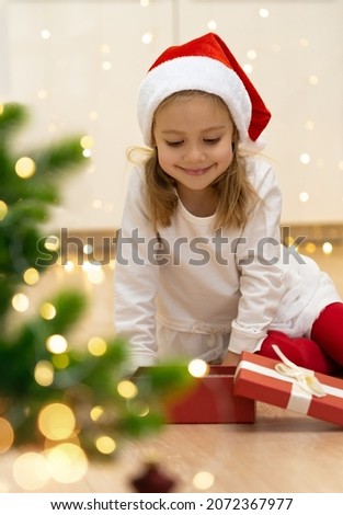 Little girl in red santa hat sits on floor with open box next to christmas tree and smiles. Happy child with New Years gift